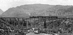 A black and white photograph of a small steam-powered train crossing the bridge, seen from upstream. There is a house along the creek at the bottom.