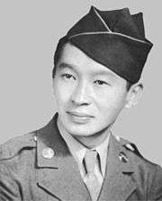 Head of a man with a subdued smile wearing a garrison cap tilted over his left ear and a military jacket over a shirt and tie.