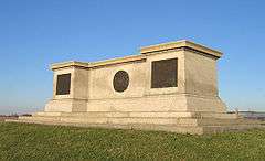 A large monument of off-white stone. Its shape resembles a long, flat pedestal. It sits atop a grassy knoll with a bright blue sky overhead.