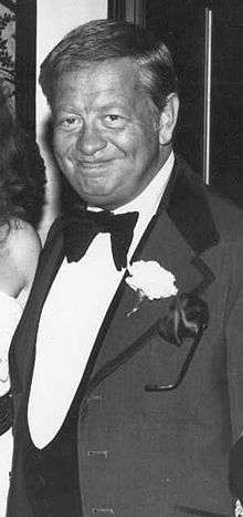 Black and white image of a man in a suit and bowtie, with a white flower pinned on his lapel