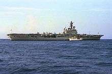 Colour photograph of a grey-painted aircraft carrier at sea. Several white aircraft are parked on its flight deck.