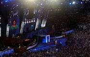 Michelle Obama speaks at a convention; her image and name are projected on a huge screen behind her. The large audience waves vertical blue signs.