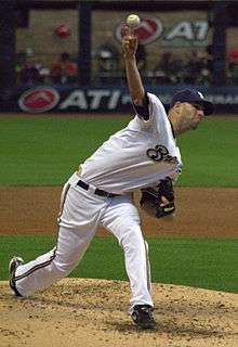 A man wearing a white baseball uniform with "Brewers" written across the chest stands on the pitcher's mound having just thrown a ball with his right hand