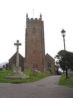 Reddish stone building with square tower. In the foreground are a cross and lamppost.