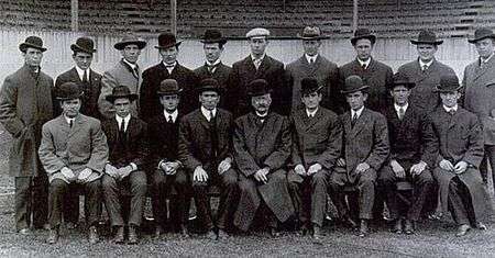 Black and white formal portrait of 10 young men standing and 9 seated in front of them. Most wearing formal hats, overcoats and suits.