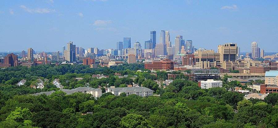 The Minneapolis skyline rises to its highest point at the center of the image, with the three tallest buildings standing out against a clear blue sky. Before the skyline are trees, university buildings, and residential complexes.