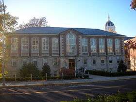 A tall brick building with a hipped roof, central pavilion and stone foundation seen from across a road with the sun just outside the left of the image.