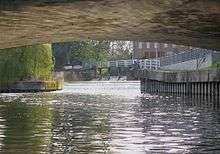 Photograph taken from centre of river underneath a wide-arched bridge. View upstream towards a weir in the far distance.