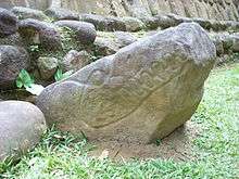 A roughly triangular boulder with the point inclined to the right. It has been carved in low relief to outline the jaws, teeth and eyes of a crocodile, with the point of the boulder representing the snout. A stonework structure rises behind the sculpture, which faces towards a grassy area to the right