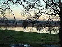 A children's play area sits amid leafless tress in the foreground. Beyond is a still body of water reflecting a cloudless sky. In the middle distance is a brown-coloured marshland, with green fields and woodlands beyond that, illuminated by a sun that is low in the sky.