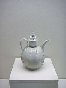 A white teapot with an almost perfectly spherical body and a large, cylindrical cap in the center which is topped with a small crown shaped embellishment. Several vertical lines are glazed into the body of the pot.