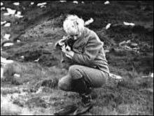 A crouched blonde woman in thick jacket, trousers, and boots, holding a small dog.