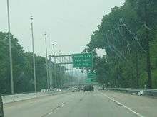 A four lane highway with a center concrete barrier containing lampposts that is surrounded by trees and has power lines running along its right side. A green overhead sign in the distance reads Morlot Ave. Fair Lawn 1/4 mile.