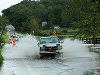 A pickup truck leaving high spray columns behind as it crosses a torrent of water crossing a two-lane road. Behind it is a line of vehicles waiting to make the same crossing