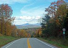 A two-lane highway in a wooded area during autumn. It drops away in the center, with a view toward a distant landscape with a body of water and mountains beyond. On the right is a sign with the number 22