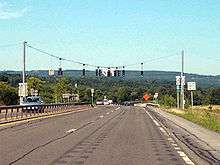 A view of the four-lane NY 5S as it approaches a traffic signal. NY 28 is accessed by turning left at the signal. In the background and distance are tree-covered mountains.