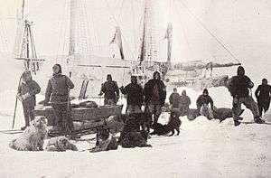  A group of men pose on the ice with dogs and sledges, with the ship's outline visible in the background