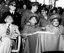 Three men seated and observing an event. The first man from the left is wearing a suit and fez, the second man is wearing a military uniform, and the third man is wearing military uniform with a cap. Behind them are three men standing, all dressed in military uniform. In the background is ab audience seated in bleachers