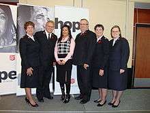 A photograph of six standing, smiling people, five of whom are wearing black uniforms with the sixth wearing a pink dress shirt