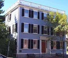 A three-story colonial-style house with a low railing around the roof. The house is gray with white trim and black shutters. There is a small portico sheltering the front door.