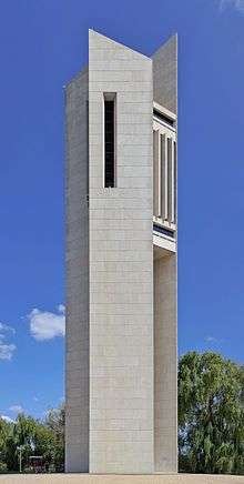 A tall modern concrete structure of three slabs, with the bells ensconced in the middle at the top, surrounded by landscaped grass and hedge.