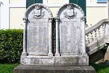 A color photo of two stone memorials, each with approximately 6 feet tall. The WW2 memorial stands on the left, with 46 names inscribed, and the WWI memorial adjoins it on the right, with 42 names inscribed.