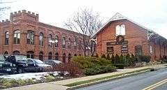 Two brick buildings viewed from their right, across a road. The one on the left has a castle-like tower on the corner; the one on the right has a decorative white wooden pointed roof. There is a parking lot in front of them on the left with some cars.