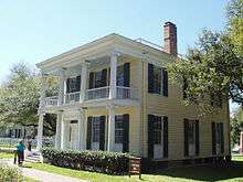 A gambrel-roofed house with large windows, gracious wrap-around balcony and first floor porch with Grecian columns. Sashed windows are framed by shutters.