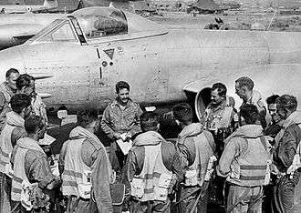 Twelve men in flying suits in a semi-circle around another man in front of a twin-engined jet aircraft