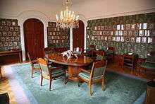 A white and green room with a wooden table in the middle, surrounded by turquoise and wooden chairs. The walls are lined with portraits and have two dark, wooden doors.