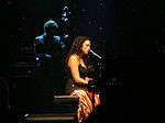 Woman in black sleeveless top and multicolored skirt sitting at a piano in a performance hall with a band behind her.