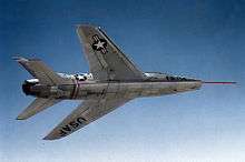 Jet aircraft in flight against blue sky; while banking away from camera, it reveals a smooth underside. A long and thin red pitot tube protrudes from the aircraft's nose