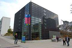 A cube-shaped building covered by a rectangular grid containing some windows, but mostly black glass. Four people are on the surrounding sidewalks and there are three banners reading "Welcome Center".