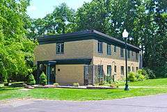 A two-story yellow brick building with a flat roof and brown trim. A metal lamppost is in front with tall trees in the back