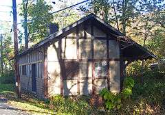 A small gold building with brown stripes and a pointed roof with a chimney on back. There is some graffiti spray-painted on it.