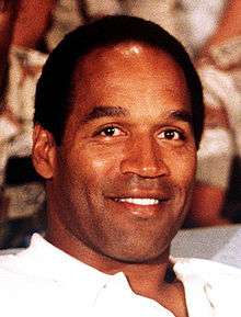 A black man smiling at a camera while wearing a white shirt.