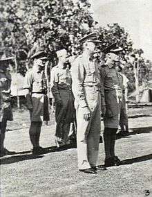 Full-length, three-quarter-angle, outdoor portrait of half a dozen or so men in light tropical military uniforms with headgear, standing to attention. Two of the men are in the foreground and the rest in a row behind them.