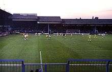 A football match in progress at a modest, traditional British football ground.