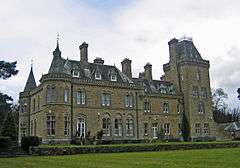 An imposing yellow-stone house in two storeys with attics seen from a slight angle. On the left are two circular turrets with conical roofs; on the right is a larger square tower with a truncated pyramidal roof.  Rising from the roofs are dormers and chimneys.