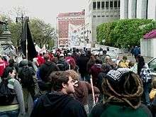 A group of people marching along a city street ahead of the camera, carrying various signs and banners. On their right is a building with tall, smooth stone facades; on the left a more ornate stone stairway and streetlamp