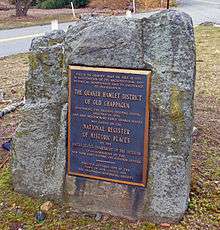 A square gray rock with a plaque on it commemorating the addition of the historic district to the National Register of Historic Places