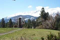 Photograph of Old Chief Joseph's gravesite, with a stone pillar on top of a hillock and the Wallowa Mountains rising in the background.