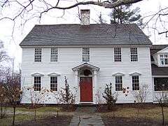 A white wooden house with no shutters, roof, and central chimney, and trees around