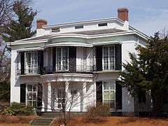 A white two story house with black shutters. Windows on both floors extend from floor to ceiling. The central portion of the house has a protruding rounded porch, supported by white fluted pillars below, and decorated on the second floor with wrought iron railings.