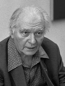 An eldery, balding man with swept back hair, wearing a suit. He faces the camera.