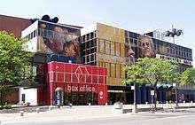 Modern building, red entrance labeled BOX OFFICE, in front of yellow and blue segments. Trees planted in front. Advertising has images of people's heads painted on building.