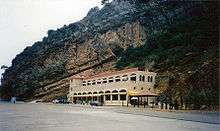 Two story stone structure built into the side of a cliff. The second floor has an outdoor balcony.