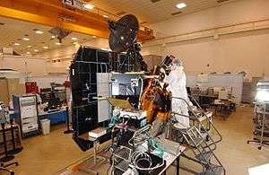 A spacecraft under construction hangs in the hallway of a laboratory as a scientist, clothed in a white protective suit, attaches scientific instruments to the spacecraft bus.