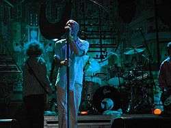 Male singer in white shirtsleeves and trousers, with a band behind him on a small stage.