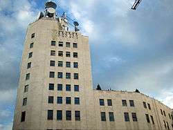 Ground-level view of a 10-storey building; the exterior has a tan hue with dark windows and a higher front side